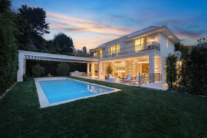 Modern luxury home exterior with illuminated windows, pool, and patio at sunset.